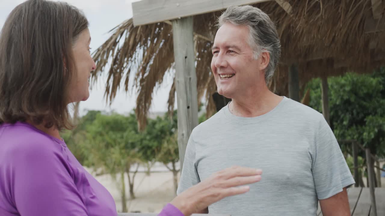um casal caucasiano feliz segurando um tapete de ioga e falando na praia, em câmara lenta.