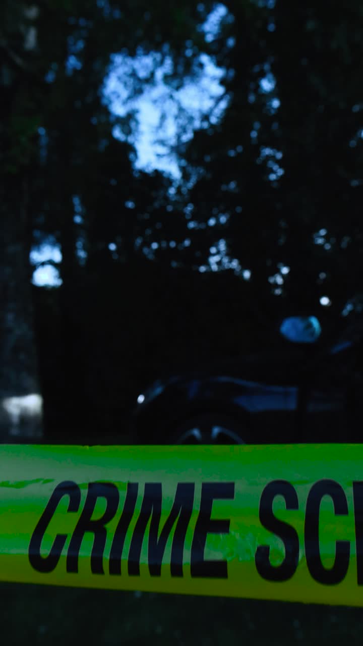Vertically filmed video of a yellow forensics police Crime Scene tape pulled in front of a crashed vehicle or car that has its lights blinking and doors opened at night time in front of dark trees