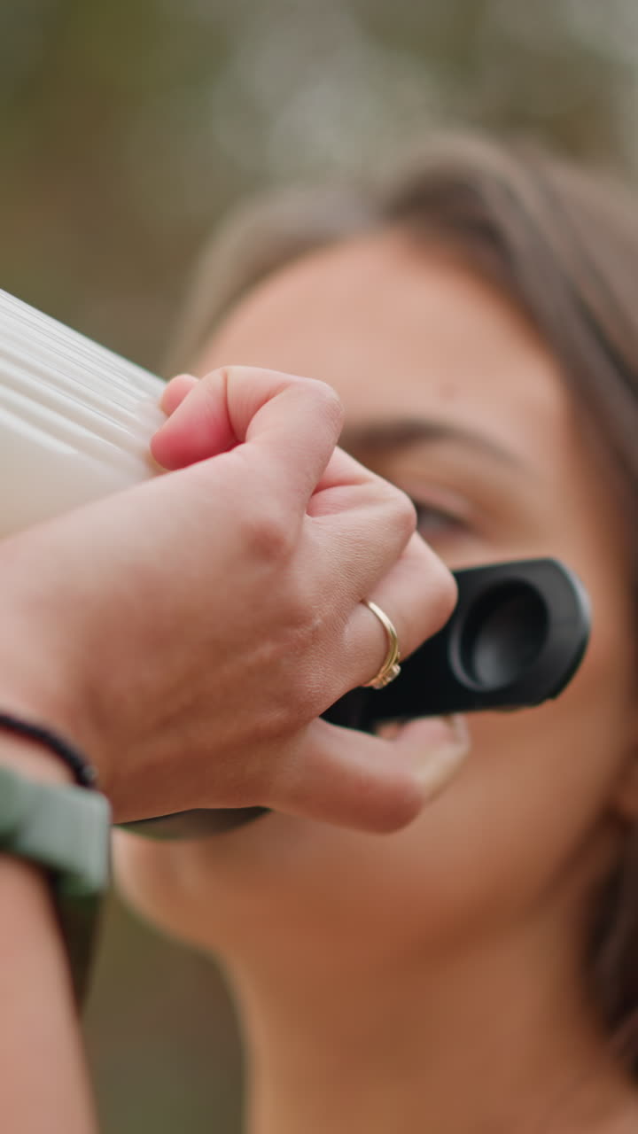 Close-up of woman drinking water from white bottle with blurred background, promoting hydration, outdoor fitness, wellness, and active lifestyle, while enjoying a refreshing drink