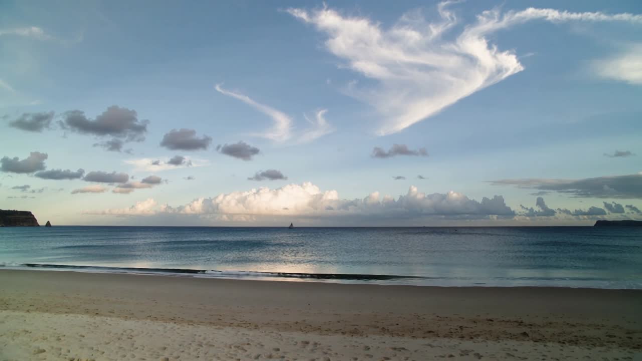 Nuvole alla deriva e onde che si infrangono mosse da una leggera brezza su una spiaggia sabbiosa con una barca a vela.