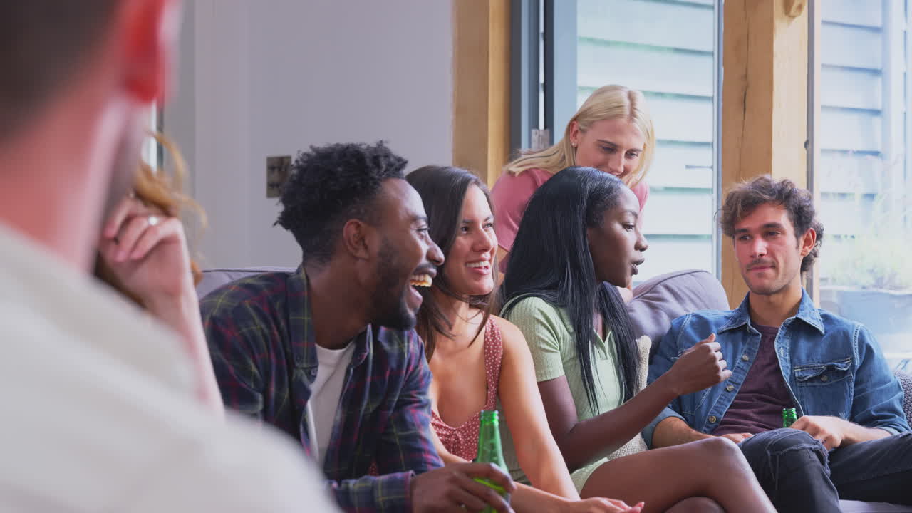 Multi-Cultural Group Of Friends Sitting On Sofas At Home Together Enjoying Drinks And Talking