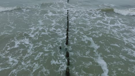 aerial birdseye view of an old wooden pier at the baltic sea coastline, overcast winter day, white sand beach, wooden poles, waves hitting shore, wide drone shot moving forward low