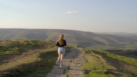 stabilised shot of young blonde woman jogging along the path on top of mam tor, castleton, peak district, england