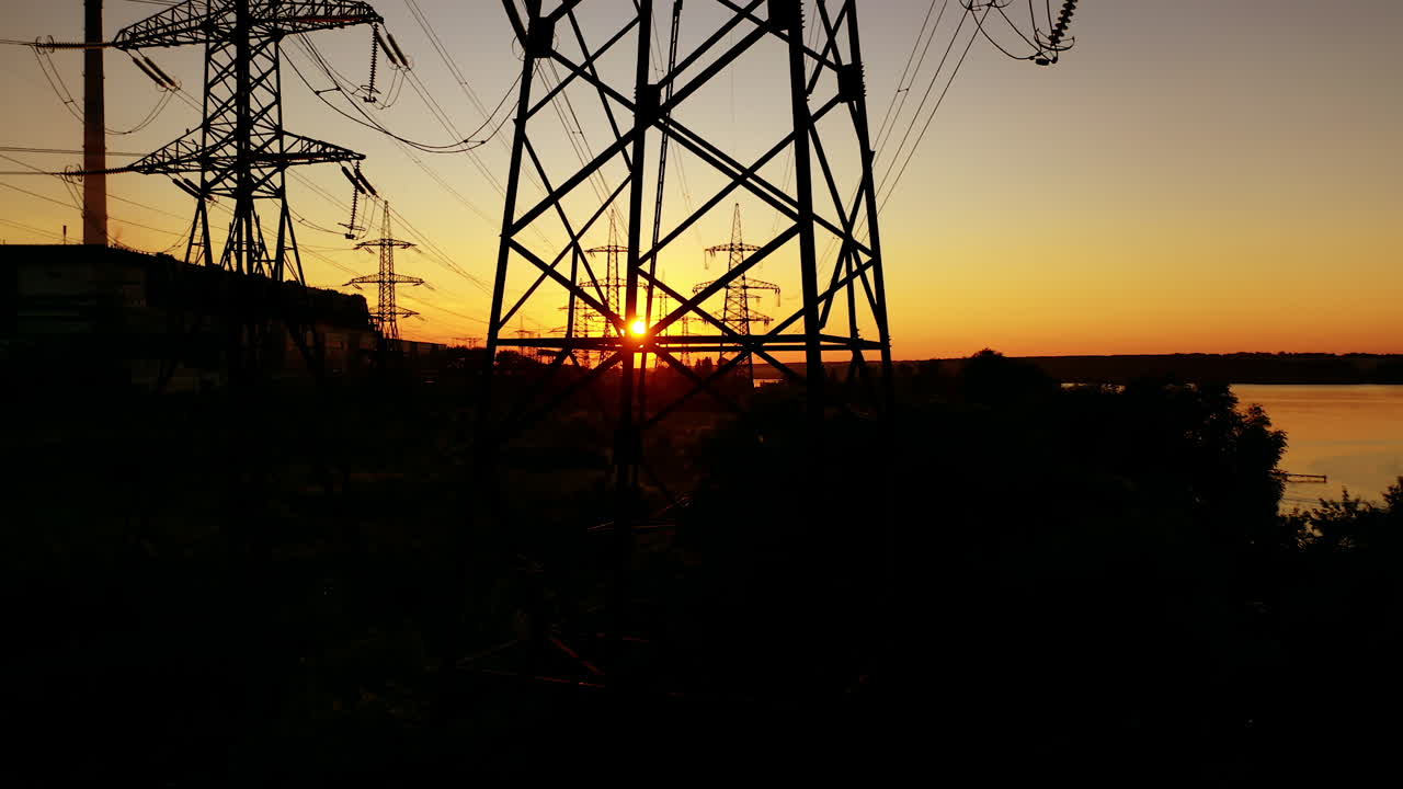 Power station near the river at sunset. Tall silhouettes of high-voltage electric lines in the evening. Electric towers on orange background of the setting sun.