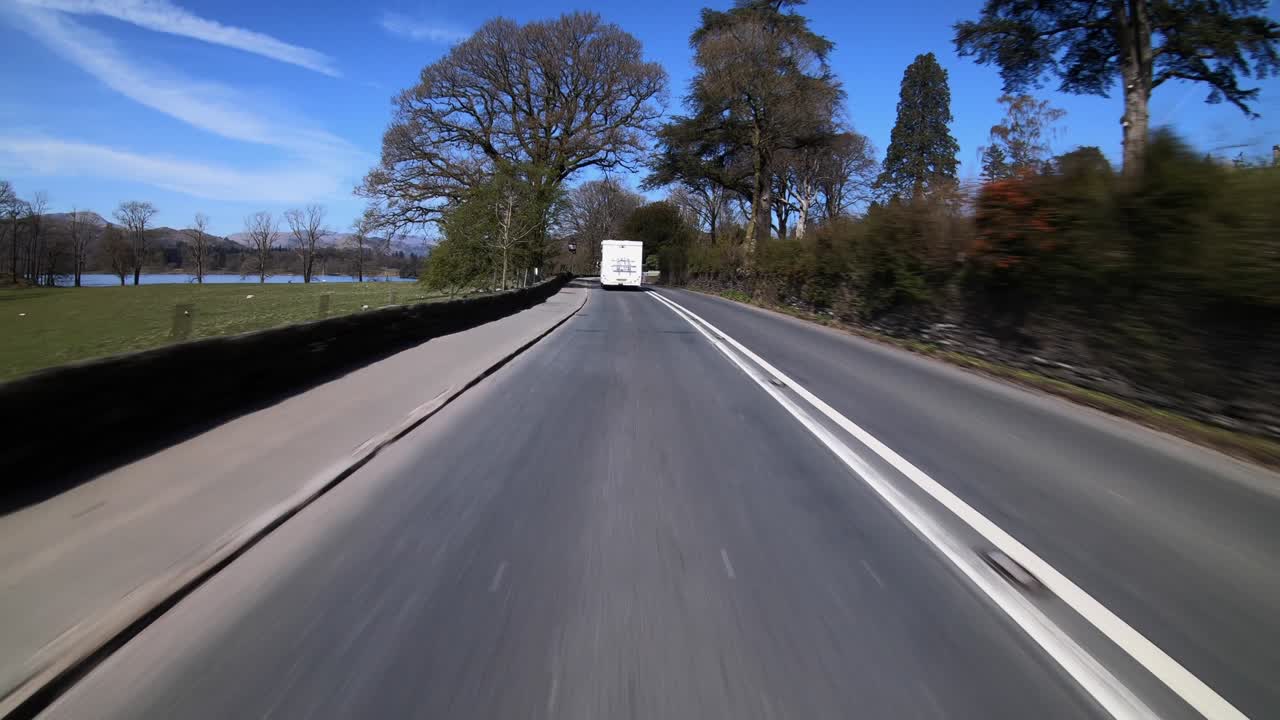 Forward POV of a Lake District road with vehicle, clear skies, and trees.