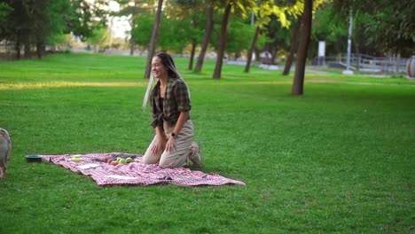 Woman-with-dreadlocks-playing-with-her-dog-on-grass,-having-picnic-outdoors-in-the-green-park-alone