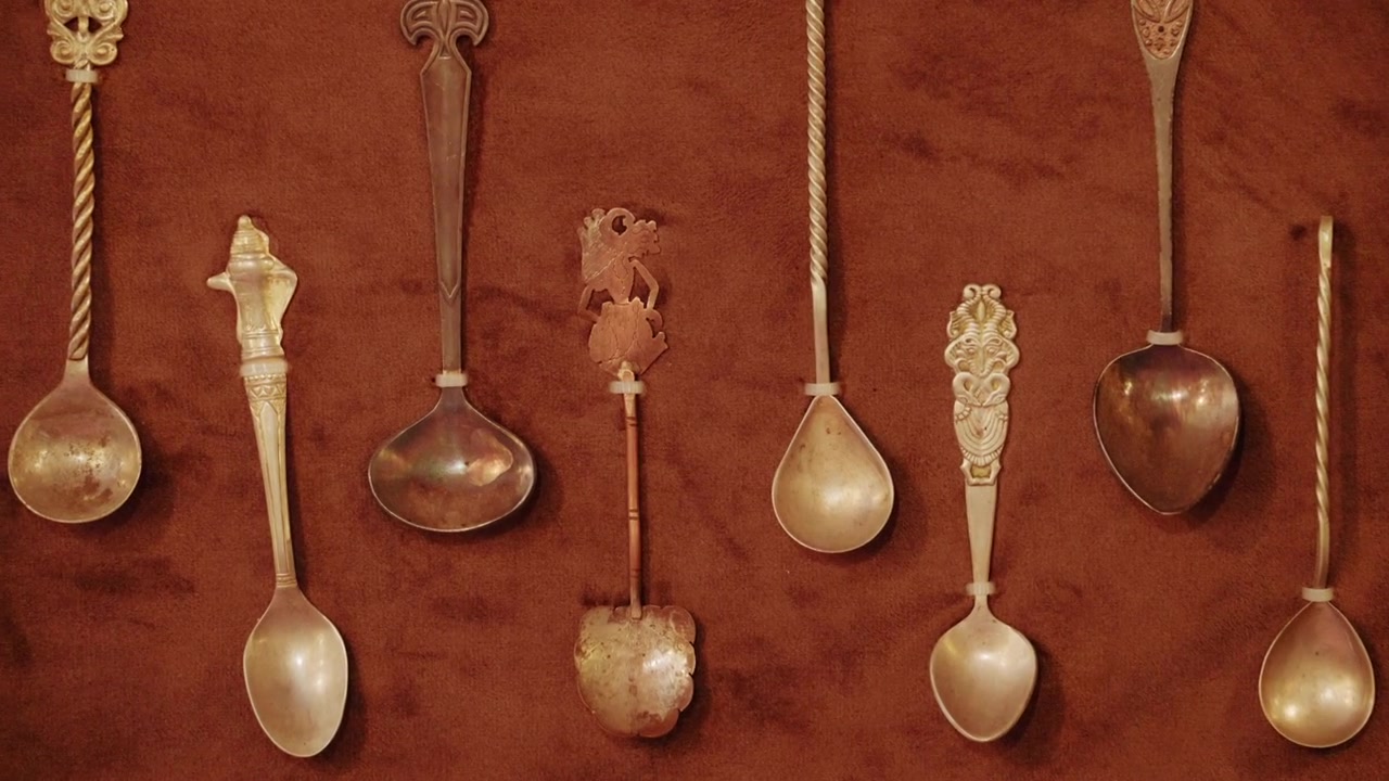 Kahverengi deri veya süet bir teşhir paneli üzerine dikey olarak monte edilmiş, altın rengi bir çerçeveyle çevrili vintage metal kaşık koleksiyonu.