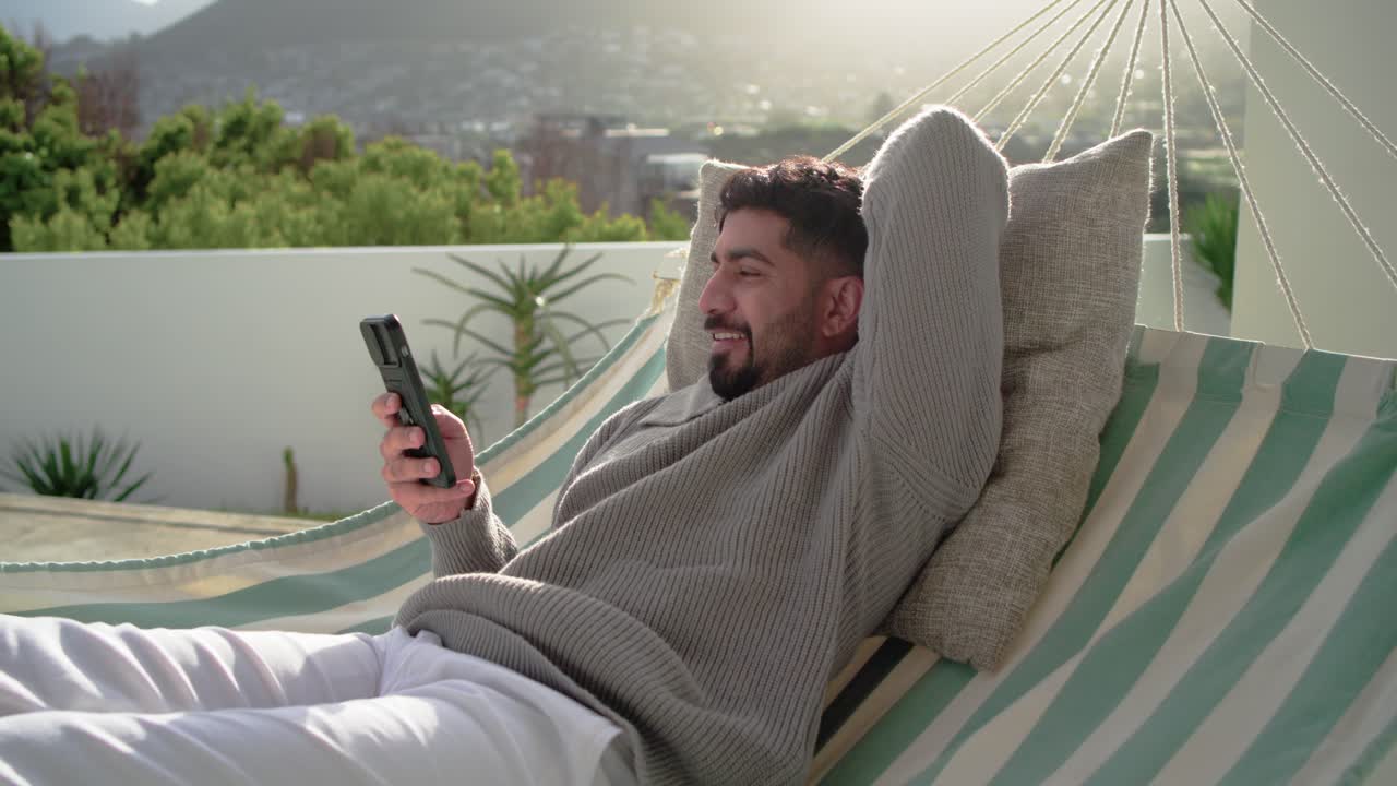 Indisk mand i 20'erne læner sig tilbage i en stribet hængekøje på en tagterrasse, læser på smartphone og ler