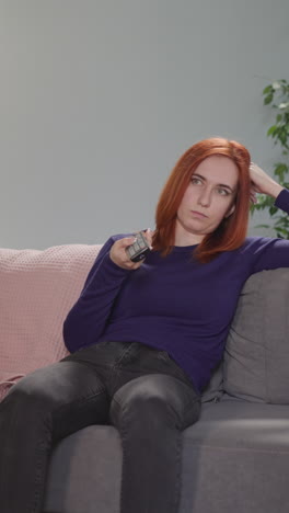 overworked housewife sits on couch changing channels on tv with remote control. red-haired woman spends evening in living room at home after work