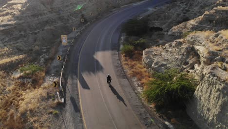 Toma-Aérea-De-Una-Moto-Conduciendo-Por-Una-Carretera-Curva-Vacía-A-Través-Del-Parque-Nacional-Hingol-En-El-Paisaje-Desértico-De-Baluchistán-Con-Destello-De-Lente