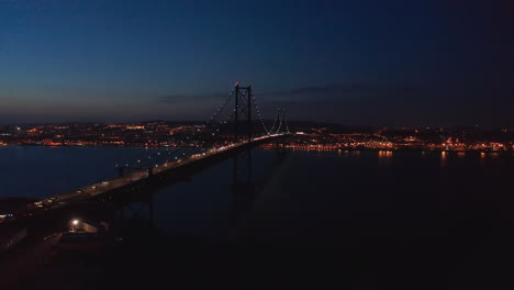 Vista-Aérea-De-La-Plataforma-Rodante-Nocturna-Del-Puente-Rojo-Ponte-25-De-Abril-Con-Tráfico-De-Automóviles-Y-El-Centro-De-La-Ciudad-De-Lisboa-Con-Luces-En-El-Fondo