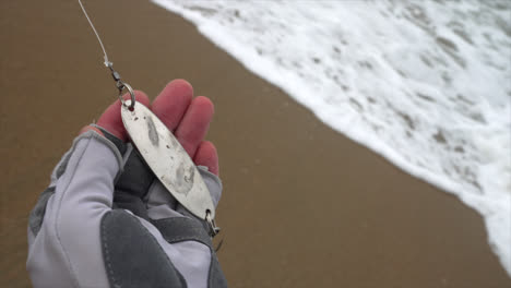 close up of hand holding silver fishing spoon lure on sandy beach