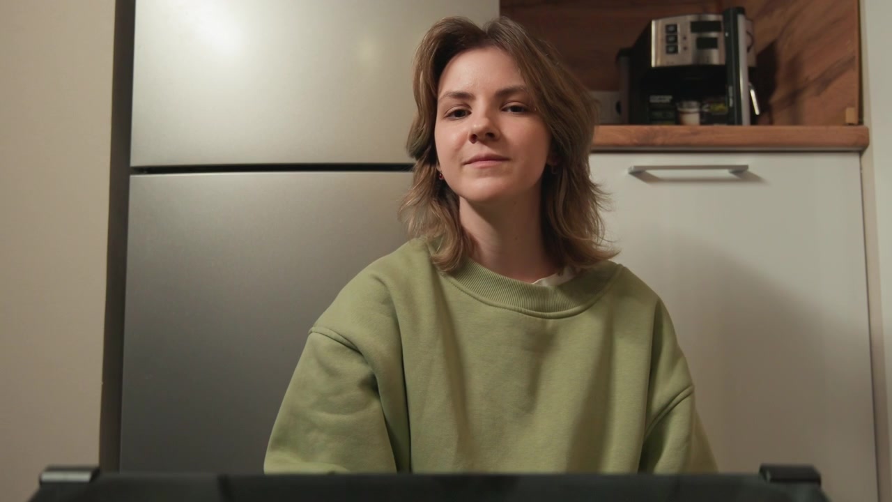 Una donna di mezza età seduta al bancone della cucina sorride verso la fotocamera con un'espressione rilassata e amichevole, in una luce calda interna e un ambiente domestico informale, con le mani fuori dall'inquadratura e un punto di vista basso a livello del bancone.