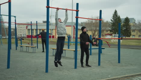 Two boys are working out on pull-up bars in an outdoor gym area, one boy is actively doing a pull-up, while the other joins him, with a man moving around in the background