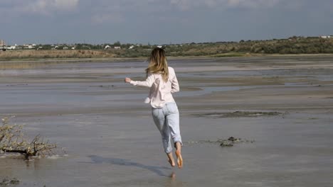 Rare-view-of-joyful-woman-in-casual-is-running-on-sandy-shallow-of-lake-at-summer