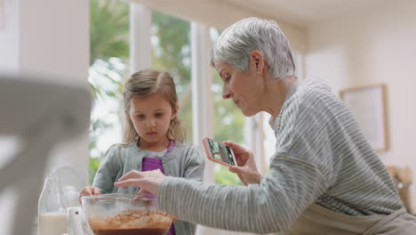 happy grandmother using smartphone taking photo of cute grandaughter in kitchen mixing ingredients for baking granny sharing weekend with grandchild on social media