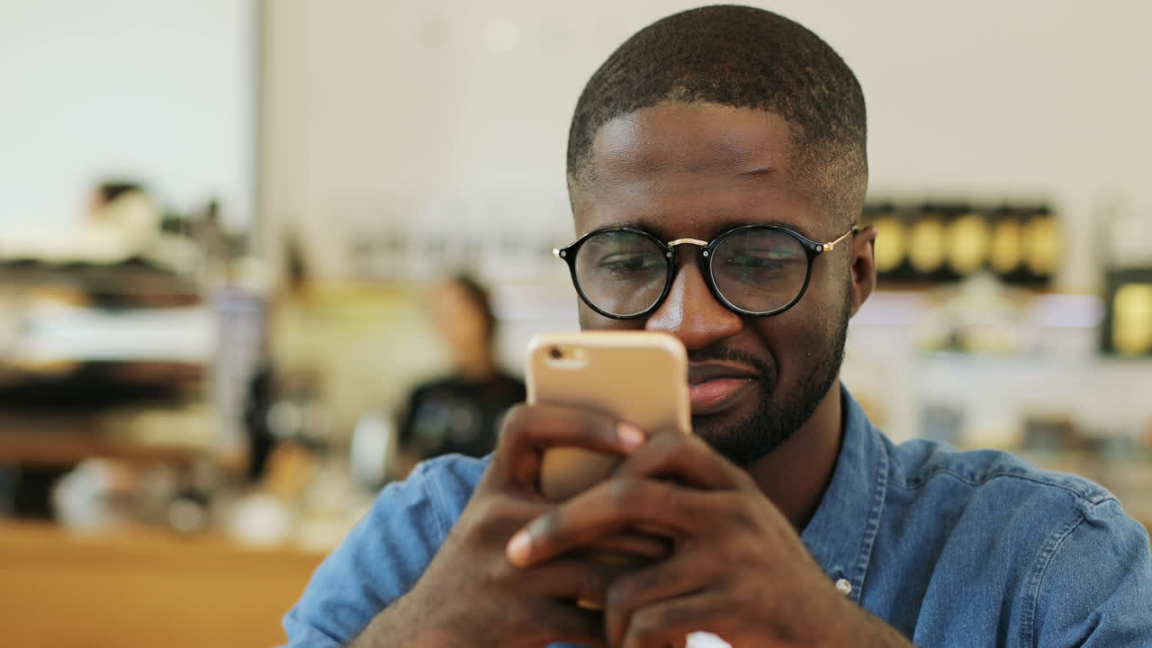 Close-up view of african american man smiling and texting on a smartphone sitting at a table in a cafe