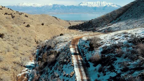 a dirt road along a canyon leading out to a valley with a lake and snow-capped mountains - aerial flyover