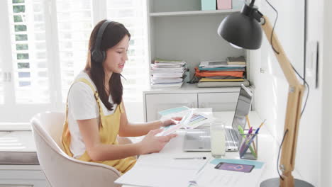 Mujer-Birracial-Con-Auriculares-Con-Videollamada-Portátil-Sentada-En-El-Escritorio-Trabajando-En-Casa,-Cámara-Lenta