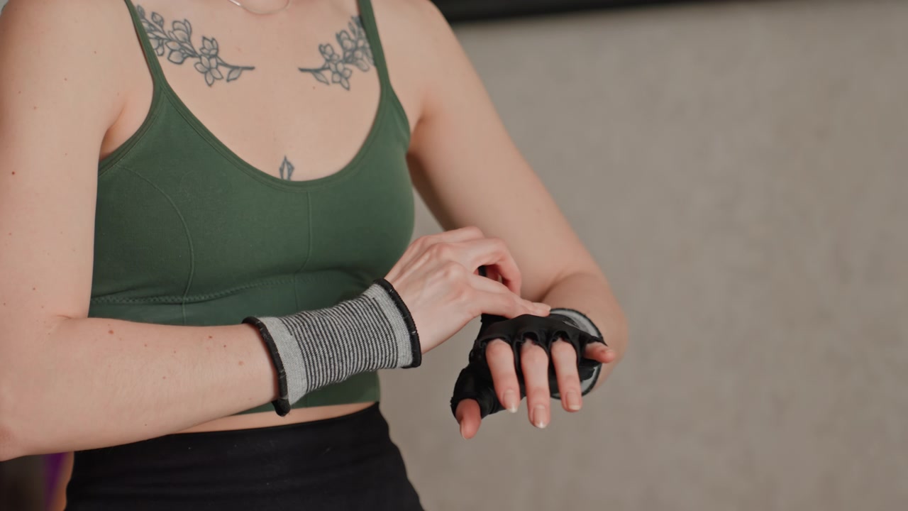 Fokus auf Heimfitness, tätowierte Frau in grünem Tanktop und schwarzen Leggings sitzt drinnen und zieht Handgelenkstützhandschuhe an, konzentrierte, ruhige Vorbereitung auf das Krafttraining, Nahaufnahme