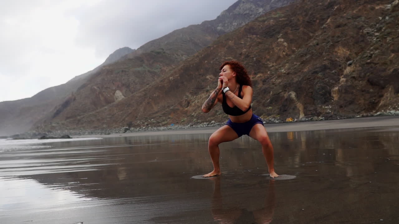 Slow motion captures a young, slender woman engaging in squats and jumps for her training by the ocean's shore