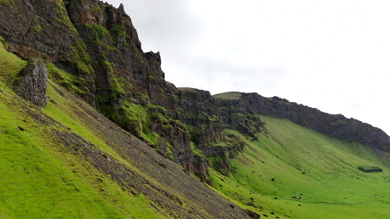 Aerial view of vibrant green fields stretching below steep moss-covered cliffs in Suðurland, Iceland, with winding streams and dramatic shadows shaping the vast volcanic landscape