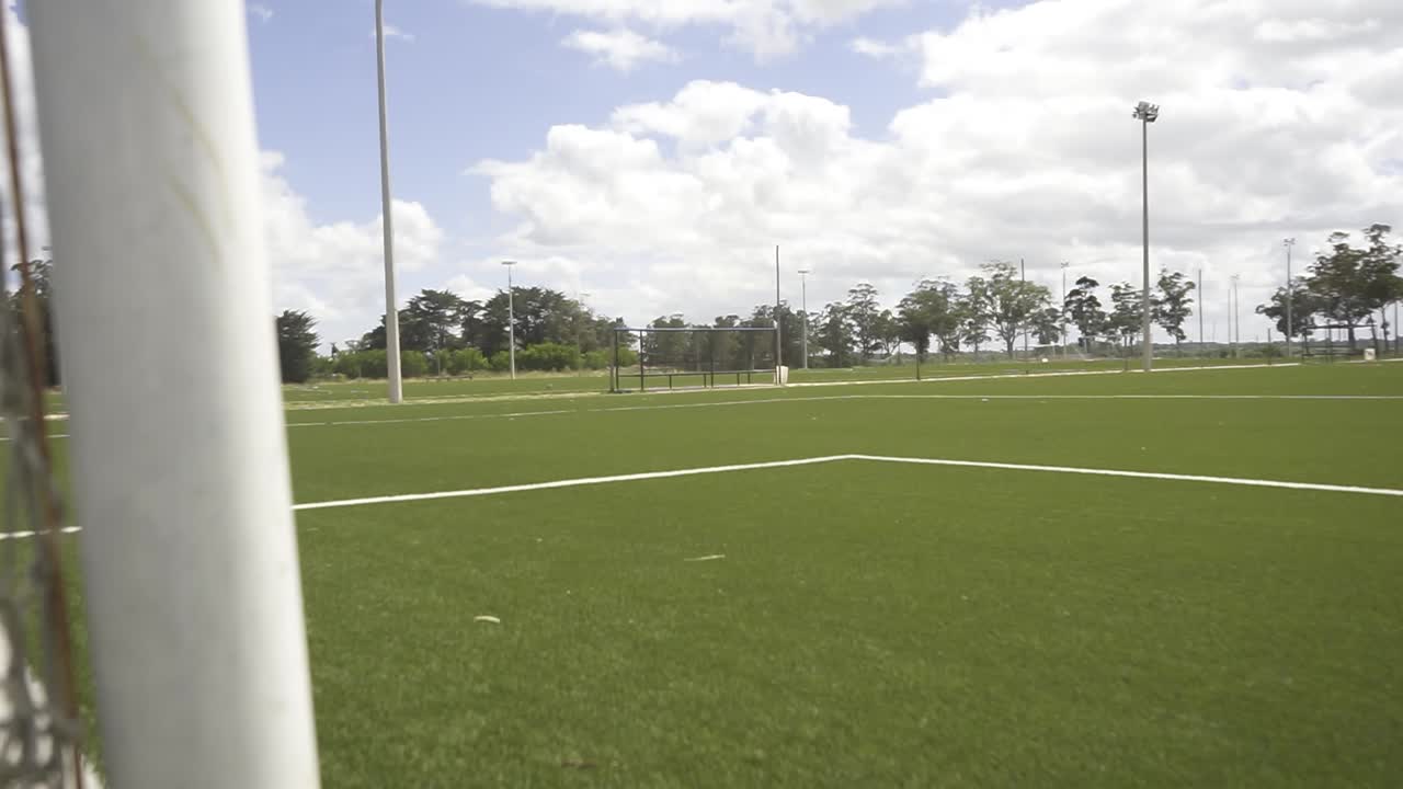 Close-up de rede de futebol revelando gramado verde, capturando cenário de estádio vazio com céu ensolarado e desfoque suave no fundo