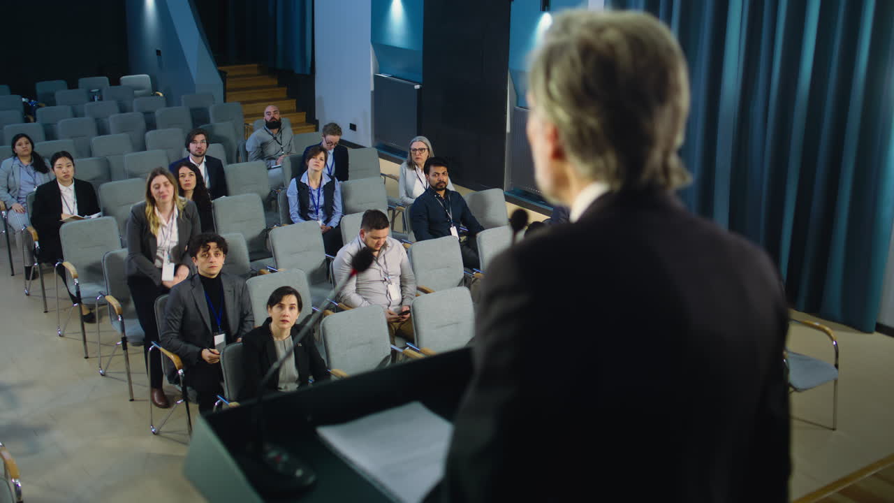vista trasera del oficial de prensa masculino pronunciando un discurso desde el escenario, dando una entrevista para la televisión o los medios de comunicación. los periodistas levantan las manos y hacen preguntas durante la campaña de prensa en la sala de conferencias.