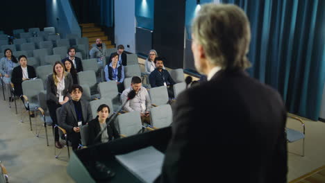 Back-view-of-male-press-officer-pronouncing-speech-from-the-stage,-giving-interview-for-television-or-media.-Journalists-raise-hands-and-ask-questions-during-press-campaign-in-the-conference-hall.