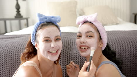 two young biracial female friends enjoy a skincare routine, wearing facial masks and headbands