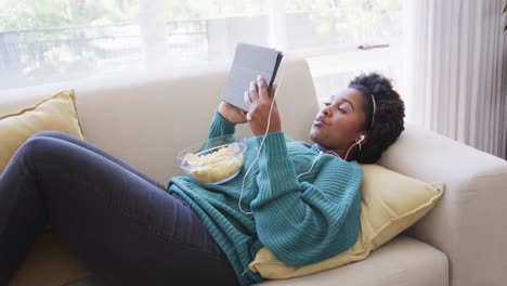 happy african american woman lying on couch in earphones, using tablet and eating chips, slow motion