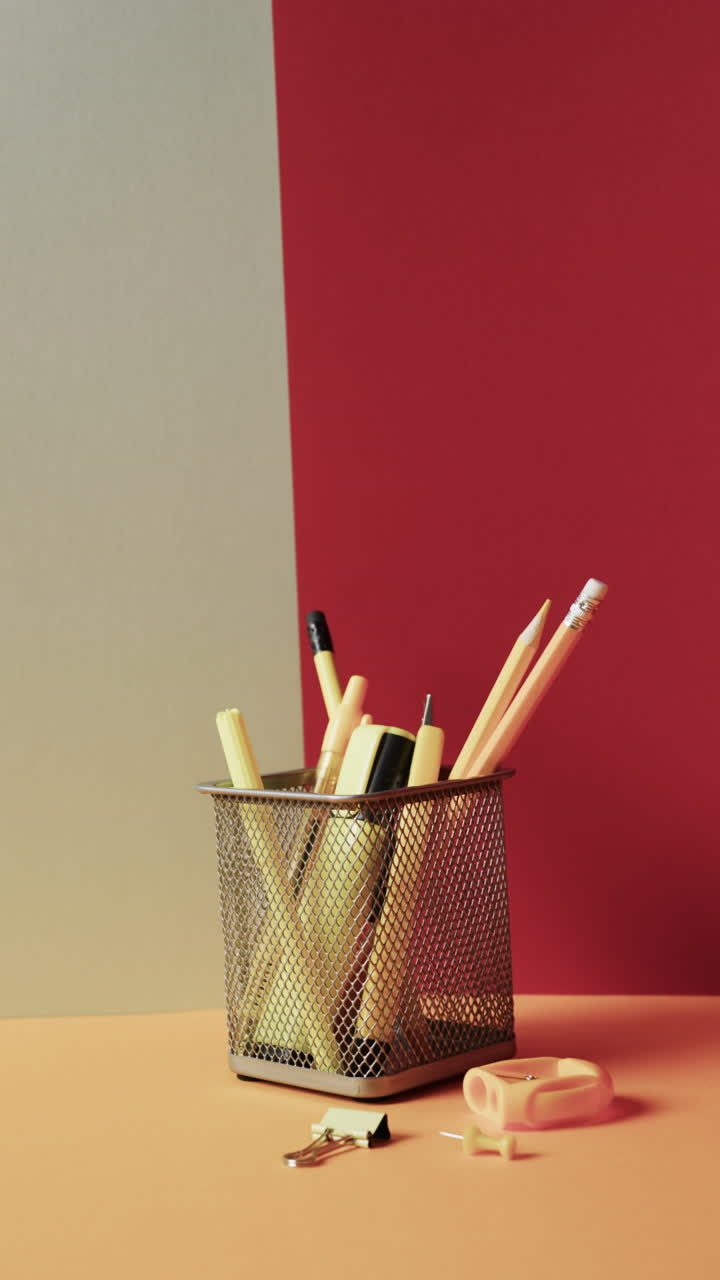 Vertical video of close up of school stationery in cup on grey and red background, in slow motion