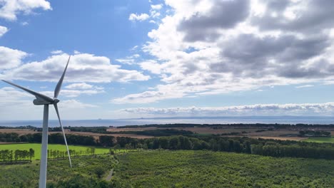 Aerial camera pans across lush green landscape, revealing a large wind turbine under partly cloudy skies. Bright daylight highlights rolling fields and distant coastline