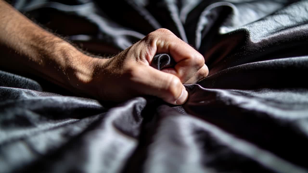 In primo piano, mano destra maschile che pizzica, tira e attorciglia un lenzuolo di raso sul letto, testandone la consistenza.