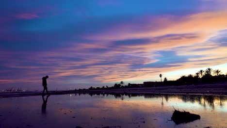 the silhouette of a man who dubiously walks and l slowly skirting the water in a beautiful sunset, the figure is reflected on the water of a lake located on the beach with awesome sky