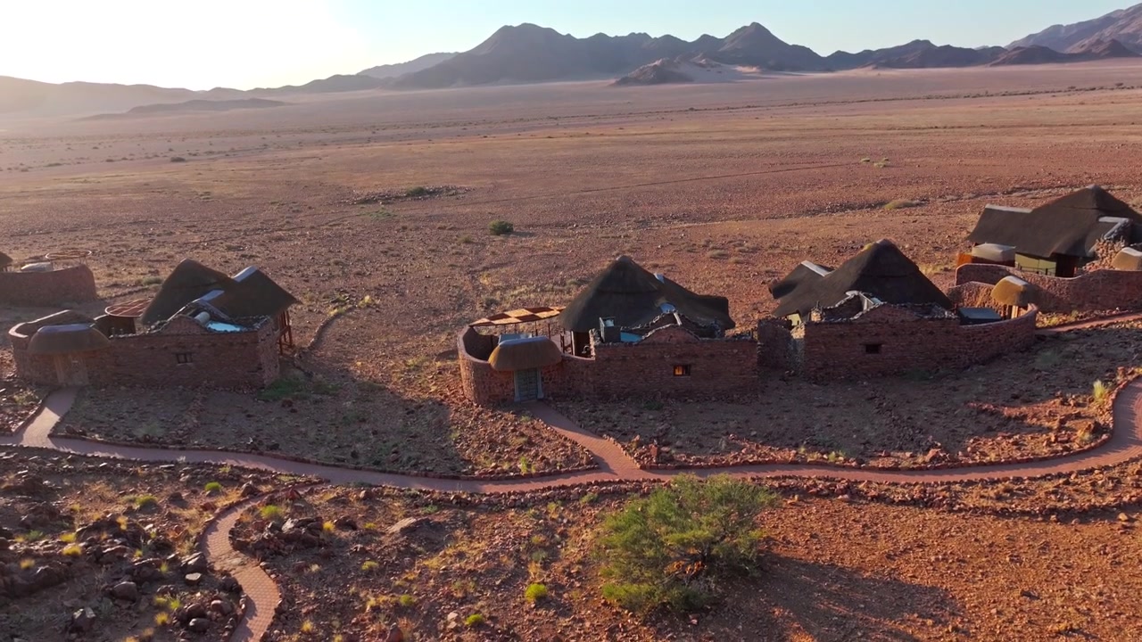 Luftaufnahme einer Wüstenlodge, eingebettet unter roten Bergen in den weiten Ebenen der Namib. Das abgelegene Öko-Retreat fügt sich in die rote Wüstenlandschaft unter weitem Himmel und fernen Bergen ein.