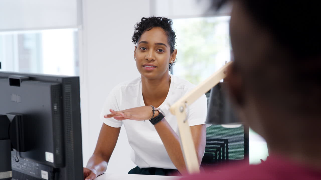 Two millennial coworkers working at computers in the office of a creative business talking to each other, selective focus