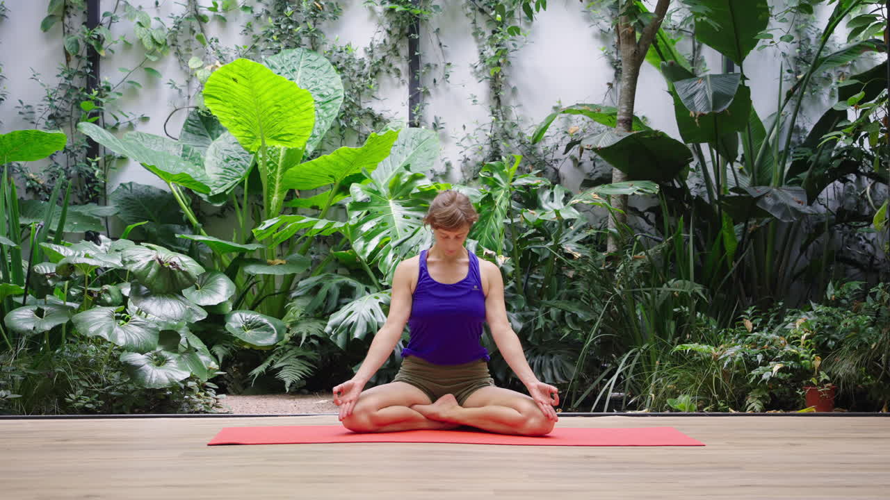 Mulher Meditando em Posição de Lótus no Tapete de Yoga Cercada por Plantas Verdes