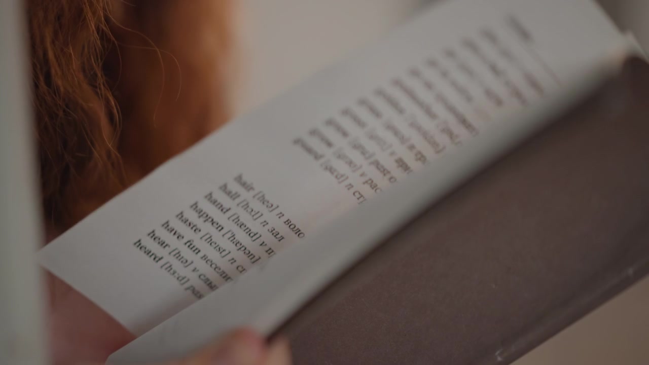 Side close-up shows hands supporting softcover book by spine and cover edge, with slight lifting motion, tilted cover, warm indoor light, shallow depth of field, calm personal reading moment
