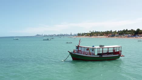 aerial drone shot of a small tour boat docked near the tropical bessa beach with skyscrapers from the coastal capital city of joao pessoa, paraiba, brazil in the background on a sunny summer day