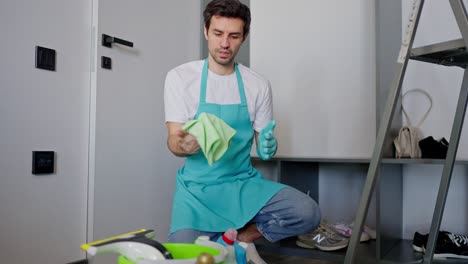 Confident-brunette-man-with-stubble-in-a-white-T-shirt-and-blue-apron-as-a-janitor-sitting-on-the-floor-among-cleaning-tools-in-a-modern-apartment