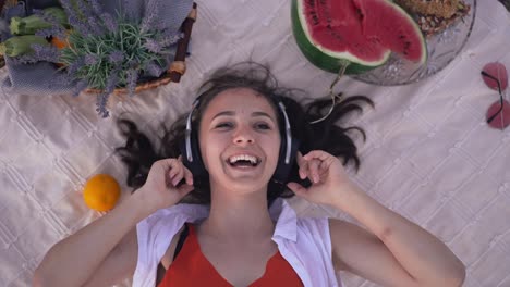 joven mujer alegre escuchando música en auriculares acostada en una manta en un picnic de verano. vista superior retrato de despreocupada feliz dama del milenio caucásica disfrutando de pasatiempo en vacaciones al aire libre.