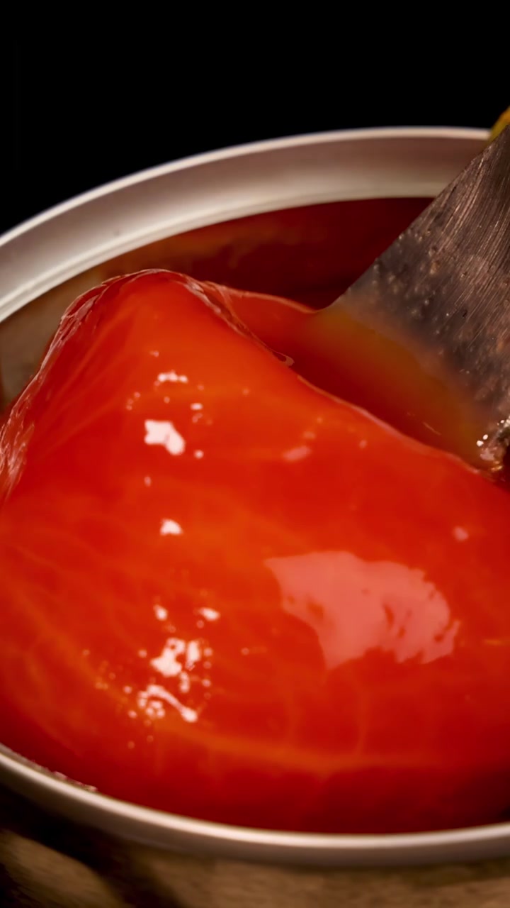 Una forchetta di metallo solleva delicatamente un pomodoro pelato intero da una lattina di succo di pomodoro sotto un'illuminazione drammatica e focalizzata con un movimento di telecamera ravvicinato