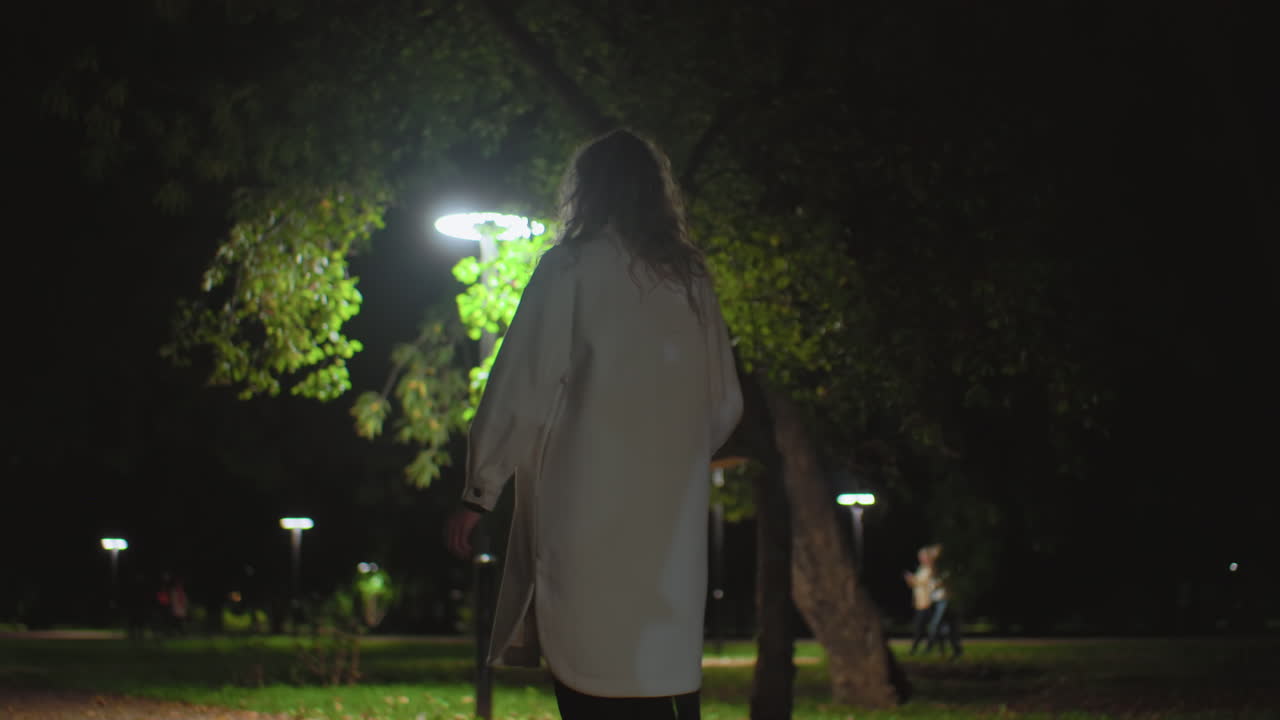 Back view of young white girl walking along dimly lit park path at night, holding umbrella, surrounded by glowing streetlights, with several blurred figures moving in distant background among trees