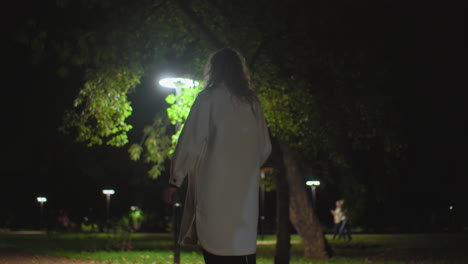 back view of young white girl walking along dimly lit park path at night, holding umbrella, surrounded by glowing streetlights, with several blurred figures moving in distant background among trees