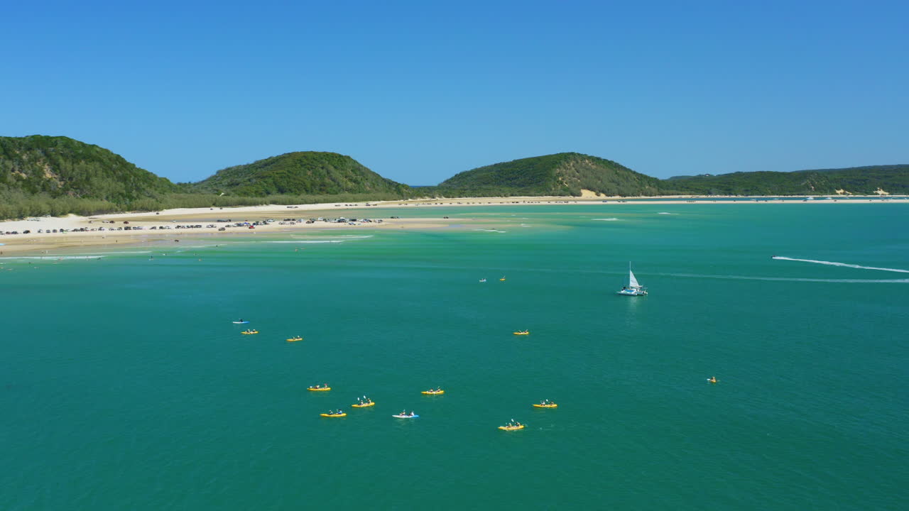 호주 이중 섬 지점의 모래 해안에서 떨어진 푸른 바다에서 카약 그룹을 패닝하는 공중 4k 드론
