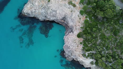 4k top-down drone footage showcasing rocky cliffs, crystal-clear blue water, vibrant reef, and green bushes along mallorca’s stunning coastline. captured on a sunny day in june 2025