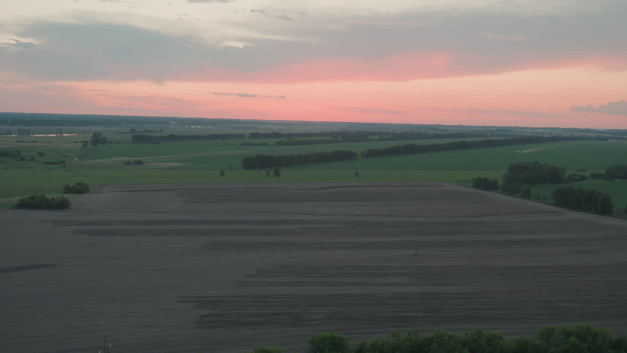 overhead drone capturing expansive harvested fields textured with soil patterns meeting green land under gentle pastel dusk sky creating serene rural panorama reaching distant horizon