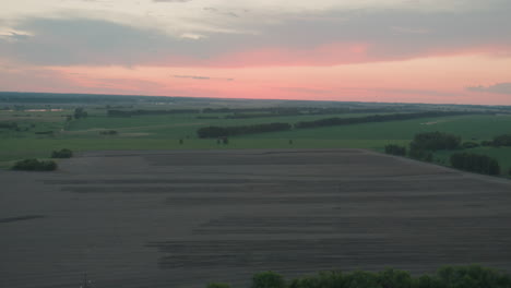overhead drone capturing expansive harvested fields textured with soil patterns meeting green land under gentle pastel dusk sky creating serene rural panorama reaching distant horizon