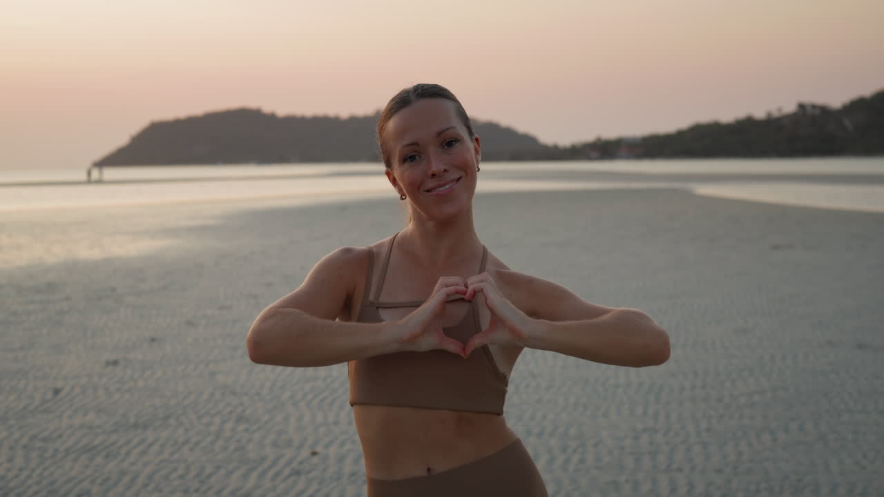 Giovane donna che forma un cuore con le mani sulla spiaggia al tramonto